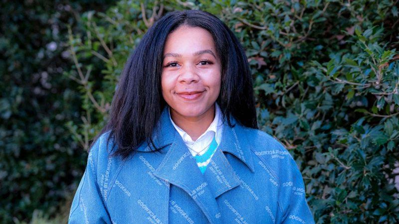 Jasmyne Yeldell standing in front of bushes with blue jacket on.